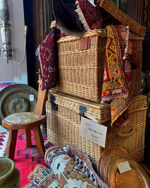 Woven baskets stacked with colorful rugs, a wooden stool, and decorative metal plates in a vintage shop setting.