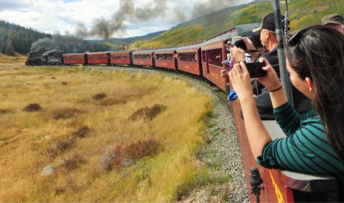 A vintage steam train travels through a scenic landscape, with passengers taking photos from the train.