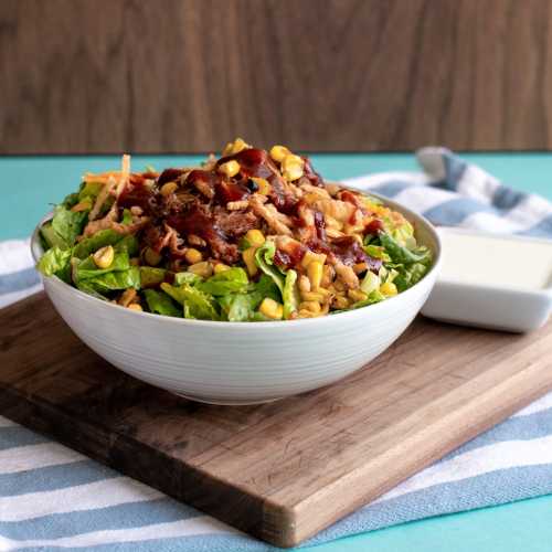 A fresh salad in a white bowl, topped with shredded chicken, corn, and drizzled with dressing, on a wooden cutting board.