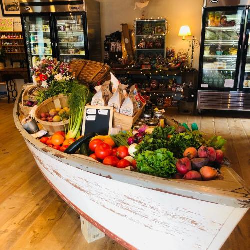 A wooden boat filled with fresh vegetables and flowers, displayed in a cozy shop with shelves and coolers in the background.