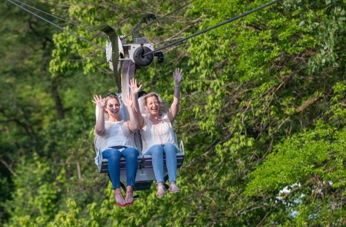 Two women joyfully wave while riding a chairlift through a lush green landscape.