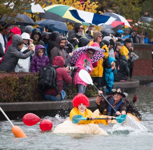 Crowd in rain gear watches two people in a pumpkin boat, one dressed as a clown, during a festive event.