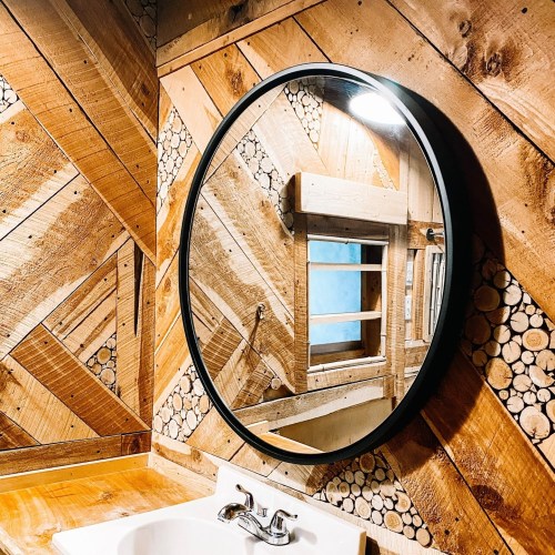 A round mirror hangs on a wooden wall with a herringbone pattern, above a white sink in a rustic bathroom setting.