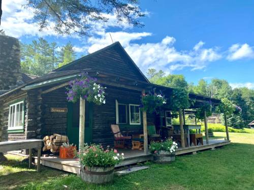 A rustic log cabin surrounded by greenery, featuring flower pots and a porch with rocking chairs.