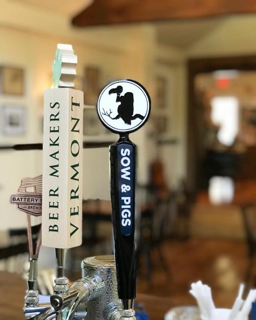 Two beer taps in a bar: one labeled "Beer Makers Vermont" and the other "Sow & Pigs." Warm, inviting atmosphere.