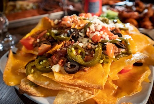 A plate of nachos topped with cheese, jalapeños, olives, tomatoes, and crumbled cheese.