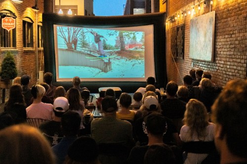 A crowd watches a film on a large screen set up outdoors in an urban setting, surrounded by brick walls and string lights.