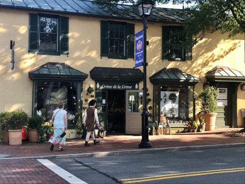 Two women walk past a charming storefront with green shutters and potted plants, featuring a sign that reads "Crème de la Crème."