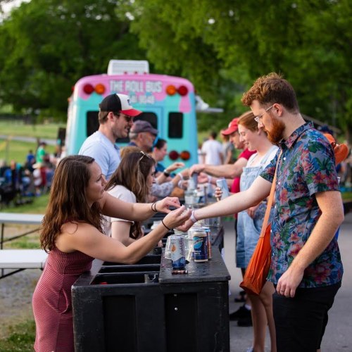 A group of people at an outdoor event exchanging drinks at a bar, with a colorful bus in the background.