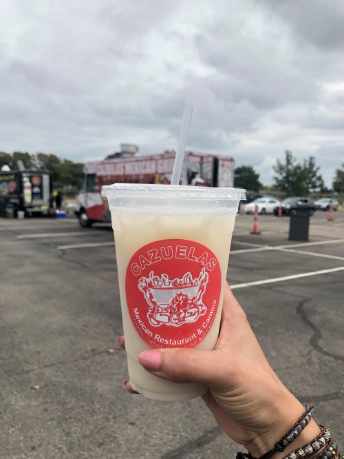 A hand holds a cup of drink with a red logo, in front of food trucks and a cloudy sky.