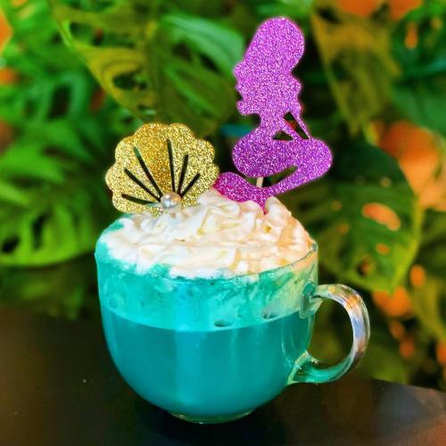 A vibrant blue drink topped with whipped cream, featuring a glittery mermaid and shell decoration.