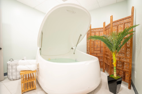 A serene spa room featuring a white floatation tank, wooden screen, towels, and a potted plant.