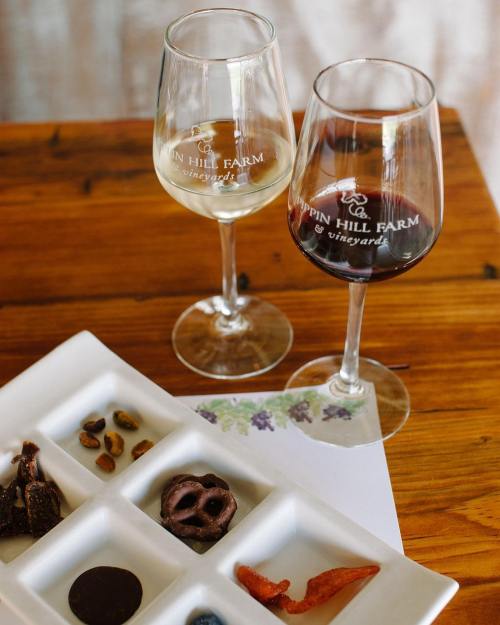 Two wine glasses, one with white wine and one with red, beside a tasting plate of chocolates and snacks on a wooden table.