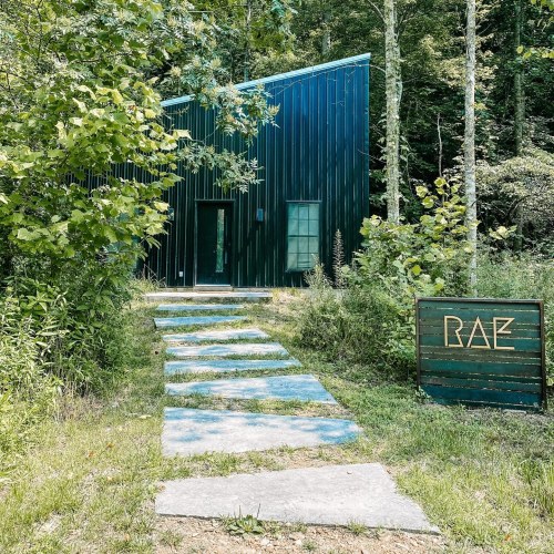 A modern green cabin surrounded by trees, with a stone pathway leading to the entrance and a sign that reads "RAE."