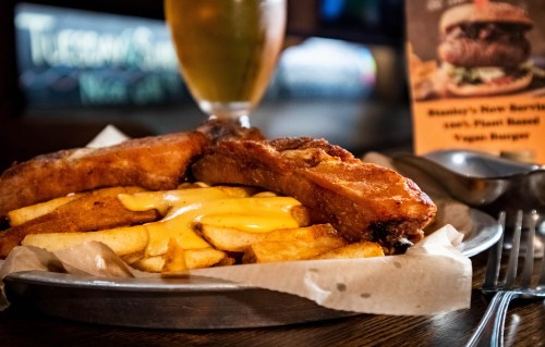 A plate of crispy fried food with cheese sauce and fries, accompanied by a drink and a menu in the background.