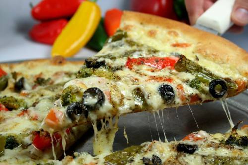 A slice of vegetable pizza with melted cheese, olives, and colorful peppers being lifted from a tray.