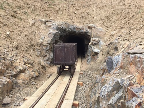 A rusty mine cart on wooden tracks leading into a dark tunnel surrounded by rocky terrain.