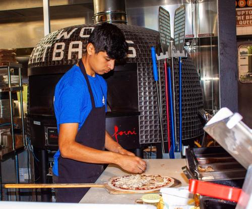 A young man in a blue shirt prepares a pizza in front of a wood-fired oven in a restaurant kitchen.