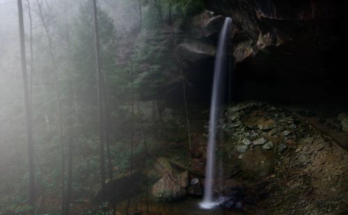 A serene waterfall cascades from a rocky overhang, surrounded by misty trees and lush greenery.