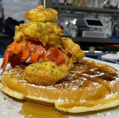 A stack of pancakes topped with shrimp and fried seafood, drizzled with syrup and garnished with herbs.
