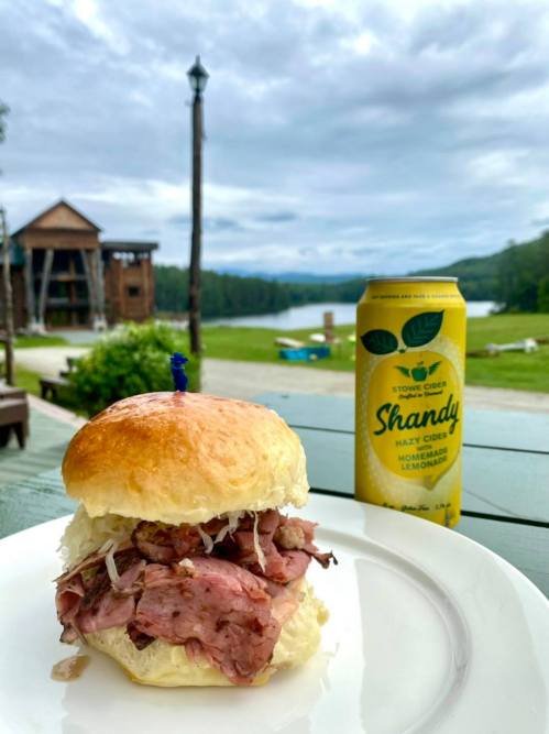 A close-up of a pulled pork sandwich on a plate, with a can of Shandy and a scenic lake view in the background.