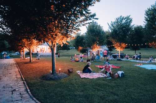 A serene outdoor gathering at dusk, with people on blankets under trees lit by warm lights.