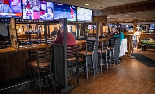 A bar area with empty stools, screens showing sports, and patrons seated at a distance behind protective barriers.