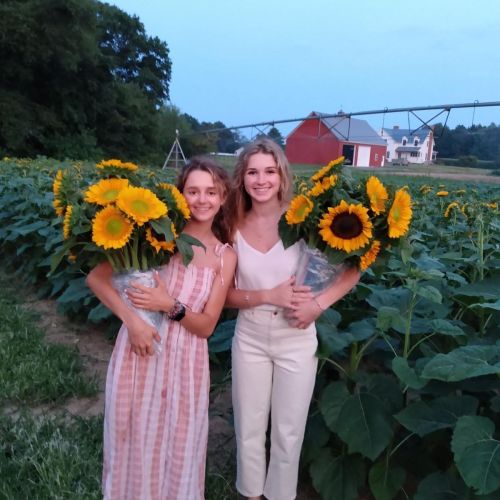 Two girls stand in a sunflower field at sunset, holding large sunflowers and smiling. A farmhouse is visible in the background.
