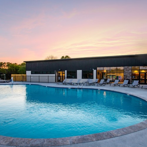 A serene swimming pool with lounge chairs, surrounded by greenery and a modern building under a colorful sunset sky.
