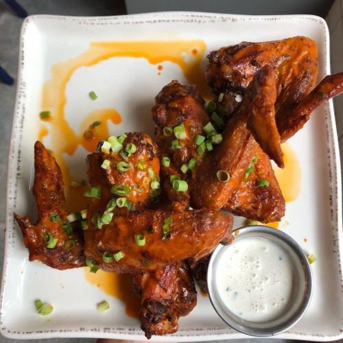 A plate of crispy chicken wings drizzled with sauce, garnished with green onions, served with a side of ranch dressing.