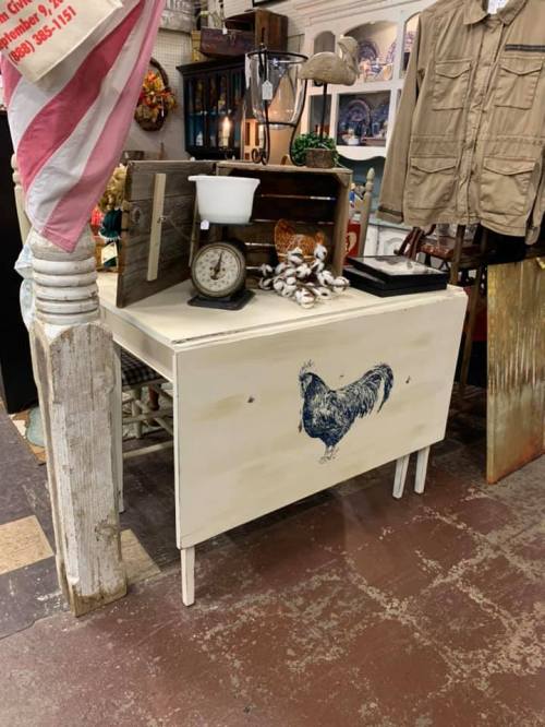A rustic table with a chicken design, a scale, and decorative items in a vintage shop setting.