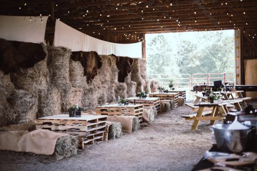A rustic barn interior with hay bales, wooden tables, and string lights, creating a cozy, inviting atmosphere.
