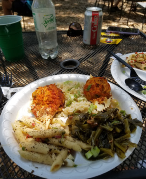 A plate with pasta, collard greens, rice, and two crab cakes, with drinks and snacks in the background.