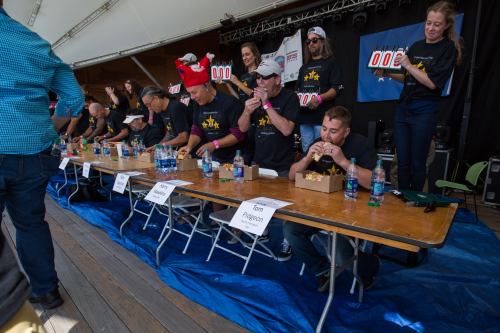 A competitive eating event with participants at tables, some eating, others cheering, under a large tent.