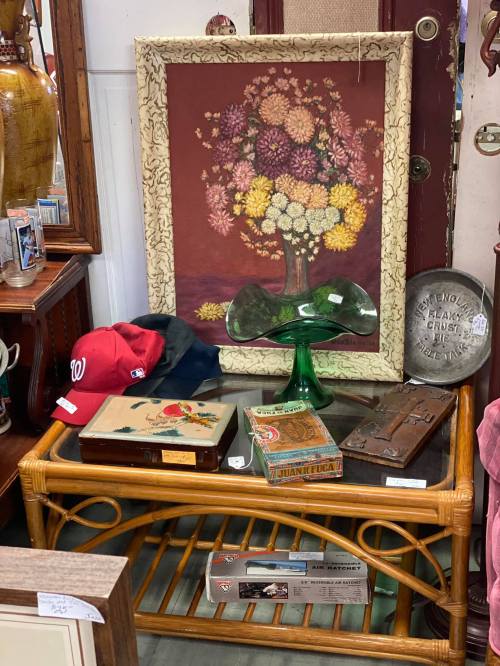 A vintage display featuring a floral painting, a green glass vase, a red baseball cap, and various antique boxes.