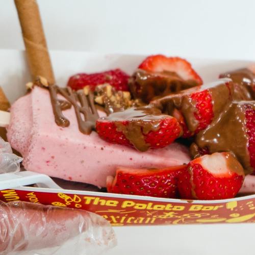 A colorful dessert featuring strawberry slices, chocolate drizzle, and a pink ice cream bar in a tray.