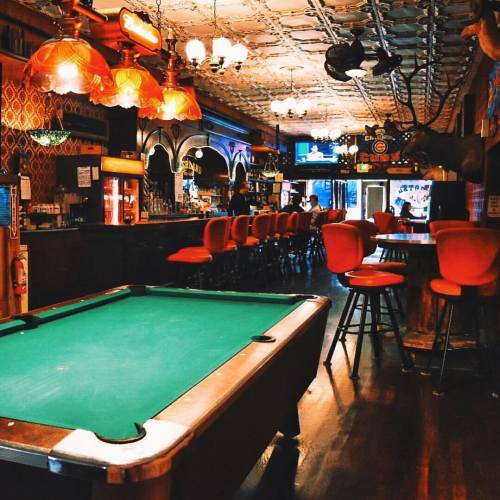 A cozy bar interior featuring a green pool table, red stools, and warm lighting, with a vintage decor ambiance.