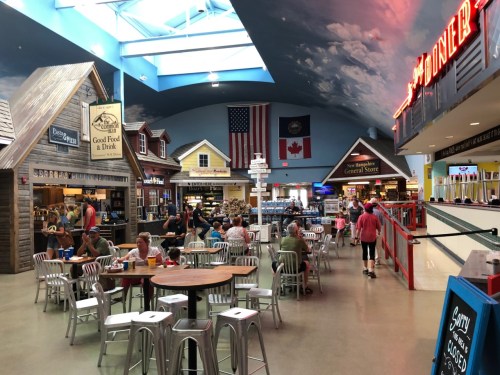 A vibrant food court with rustic decor, seating areas, and people dining under a bright skylight.