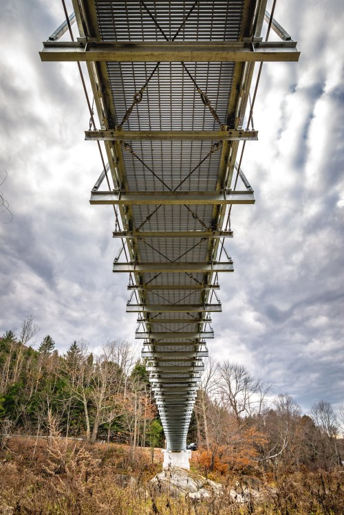 View from below a modern bridge, showcasing its steel structure against a cloudy sky and surrounding trees.