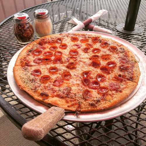 A pepperoni pizza on a metal tray, placed on a black mesh table with condiments in the background.