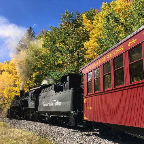 A vintage steam train travels along a scenic track surrounded by vibrant autumn foliage.