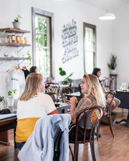 Two women sit at a table in a cozy café, engaged in conversation, with plants and decor in the background.