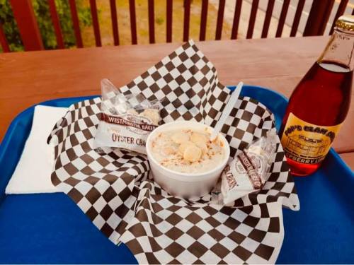 A bowl of clam chowder with oyster crackers, served on a checkered paper, alongside a bottle of soda on a blue tray.
