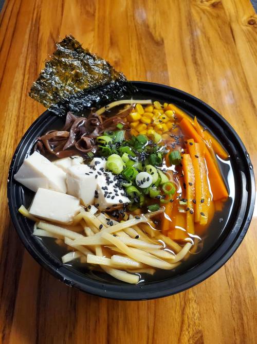 A bowl of ramen with tofu, vegetables, seaweed, and sesame seeds on a wooden table.