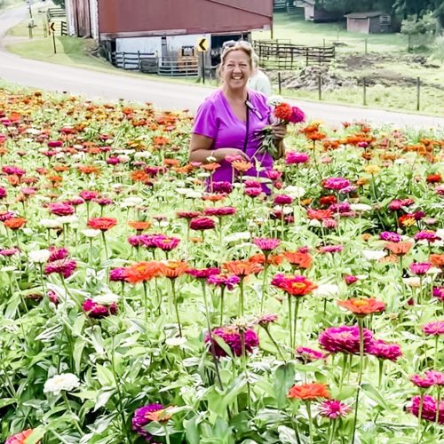 A woman in a purple dress smiles while surrounded by vibrant flowers in a field, with a rustic barn in the background.