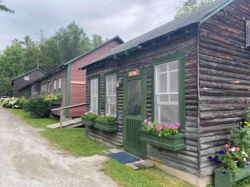 A rustic wooden cabin with green trim and flower boxes, surrounded by greenery and other similar buildings.