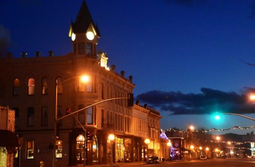 Historic buildings illuminated at night, with a clock tower and festive lights against a dark blue sky.