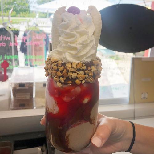 A hand holds a jar filled with a sundae topped with whipped cream, a cherry, and crushed nuts, drizzled with syrup.