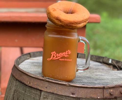 A mason jar filled with apple cider topped with a large donut, resting on a wooden barrel.