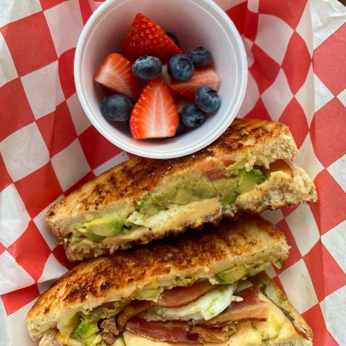 A grilled sandwich with avocado, bacon, and cheese, served with a cup of strawberries and blueberries on a checkered paper.